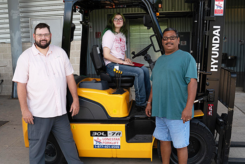 A woman sits in a forklift while two men stand nearby