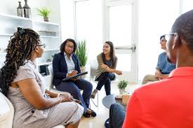 A group of smiling young adults sits indoors in a circle talking. One holds a tablet computer.