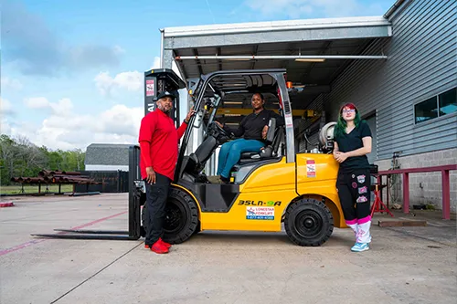 A woman sits in the cab of a forklift as two other people stand beside it